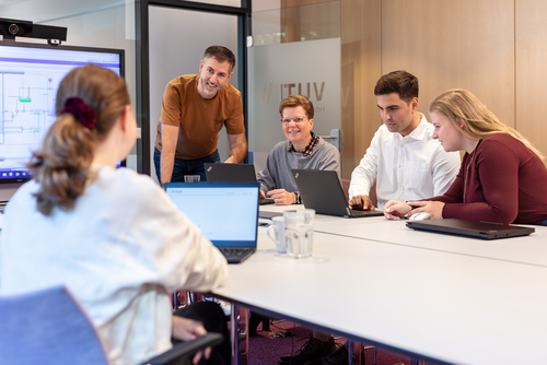 Vier Personen sitzen zusammen in einer Besprechung in einem Besprechungsraum des Unternehmens. Eine Person erklärt gerade etwas. Die anderen hören zu. Es herrscht eine freundliche und lockere Atmosphäre. 