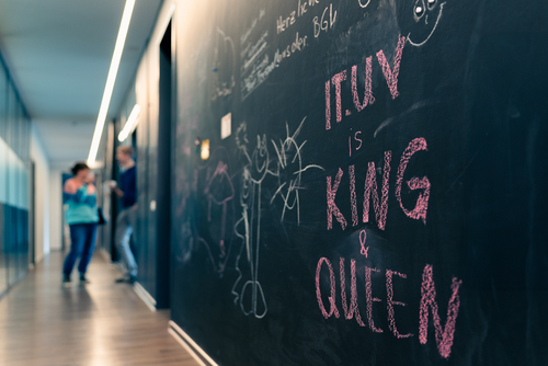 Ein langer Büroflur. Im Hintergrund stehen zwei Personen, die sich unterhalten und dabei Kaffee trinken. Sie sind nur verschwommen zu sehen. Die rechte Seite des Flurs besteht aus einer Tafelwand. Dort steht mit Kreide geschrieben „IT.UV ist KING & QUEEN“. 